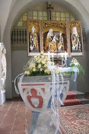 Ein Blumenstrauß in einem Steingefäß. Im Hintergrund ist das christliche Kreuz und religiöse Illustrationen zu erkennen