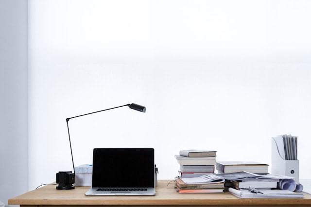 Auf dem Bild ist ein Schreibtisch zu sehen, auf welchem Bücher, ein Laptop und eine Lampe stehen